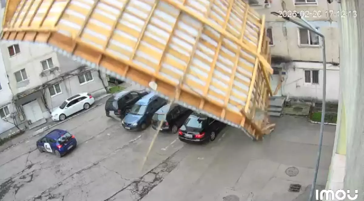 O cameră de supraveghere surprinde momentul prăbușirii unui acoperiș de pe un bloc înconjurat de mașini parcate și clădiri adiacente.