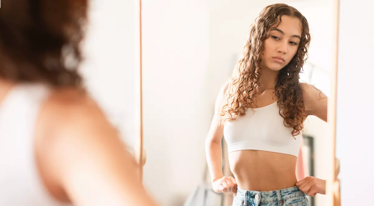 O tânără stă în fața oglinzii și își privește atent corpul, cu o expresie serioasă și concentrată. Atmosfera este una intimă, sugerând reflecție și autoevaluare a imaginii de sine.