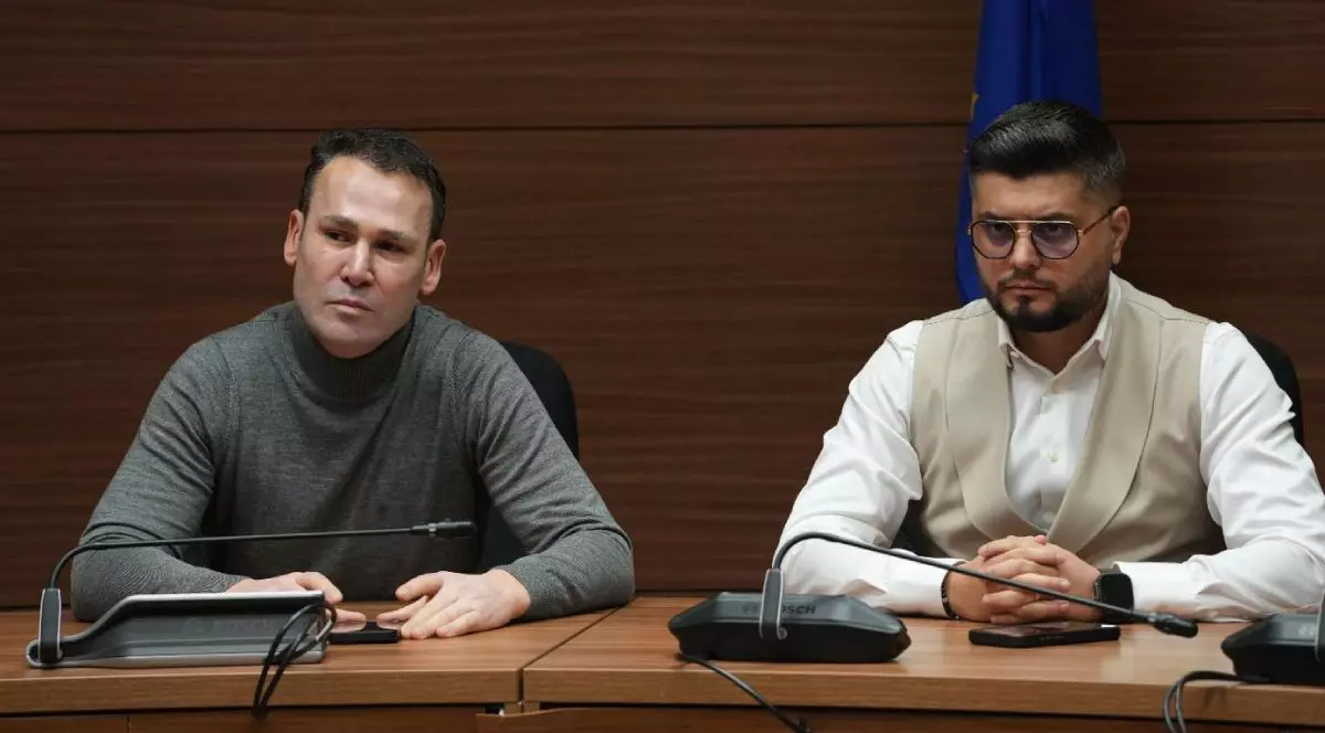 Robert Negoiță în stânga și Adrian Cocoș în dreapta, așezați la o masă de conferințe din lemn într-un cadru formal. Negoiță are părul scurt și negru și un pulover cu guler înalt gri, privește direct spre cameră. Bărbatul din dreapta, cu părul scurt și negru, ochelari și o cămașă albă sub un veston bej, privește în jos. Ambii bărbați au microfoane în fața lor. Pe peretele din spate atârnă un steag.