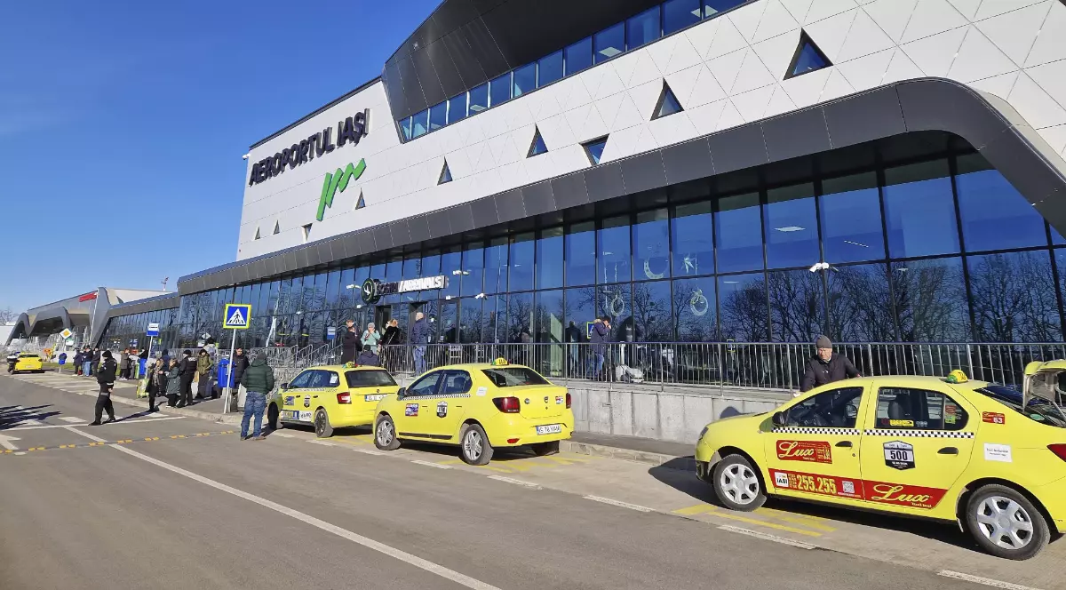 În imagine este surprins Aeroportul Internațional Iași, cu fațada modernă a terminalului și zona de sosiri, unde sunt parcate mai multe taxiuri. În fața clădirii se află pasageri și pietoni, sugerând un aeroport activ și un flux constant de călători.
