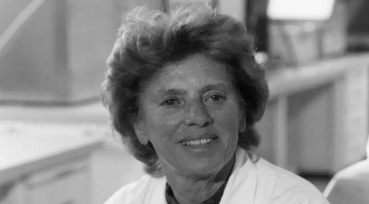 O femeie este surprinsă într-un cadru alb-negru, purtând un halat de laborator și având o expresie calmă, ușor zâmbitoare. Imaginea sugerează un mediu științific și transmite profesionalism, experiență și echilibru.