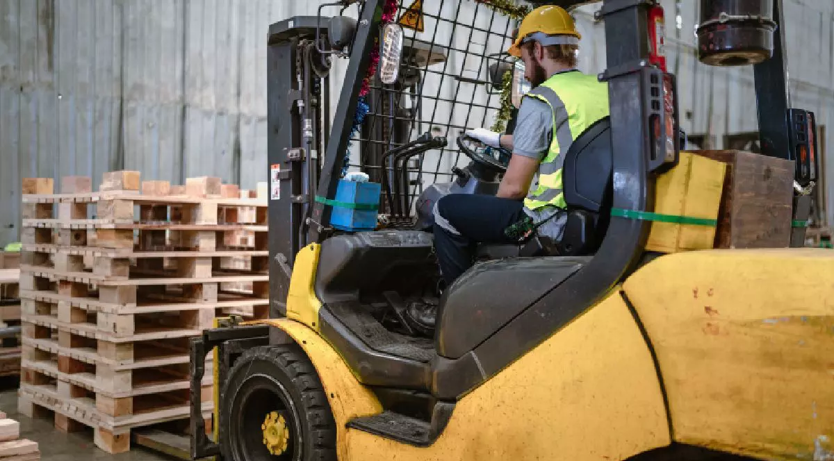 Imaginea prezintă motostivuitor galben cu un muncitor care îl operează. Bărbatul, îmbrăcat cu o vestă de siguranță galbenă, cămașă gri și pantaloni albaștri, stă așezat în cabina utilajului, care ridică mai mulți paleți de pe podea, într-un depozit.