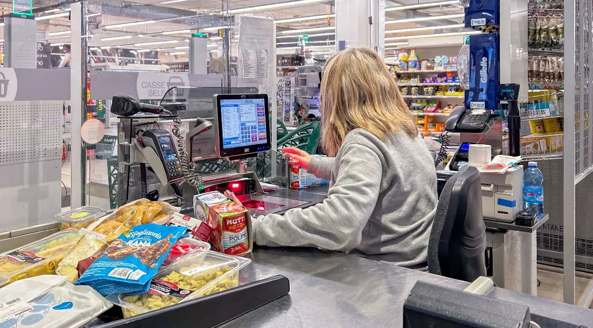 O femeie blondă îmbrăcată într-un pulover gri scanează produsele alimentare la casa de marcat a unui supermarket, înconjurată de rafturi cu produse și de un ecran digital.