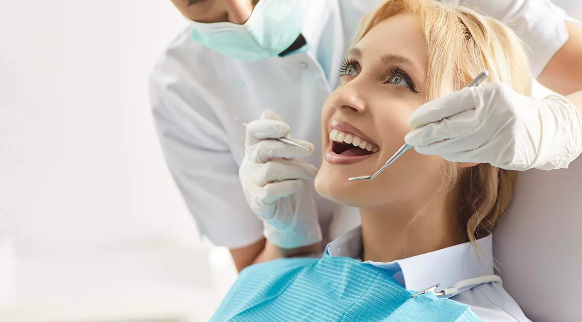 O femeie blonda sta pe scaun la stomatolog cu gura deschisa si o femeie medic stomatolog imbracata in halat alb si cu masca la gura are instrumente stomatologice in mana. Stomatologi romani, programul AP Stoma, stomatologie la stat. Foto: Shutterstock