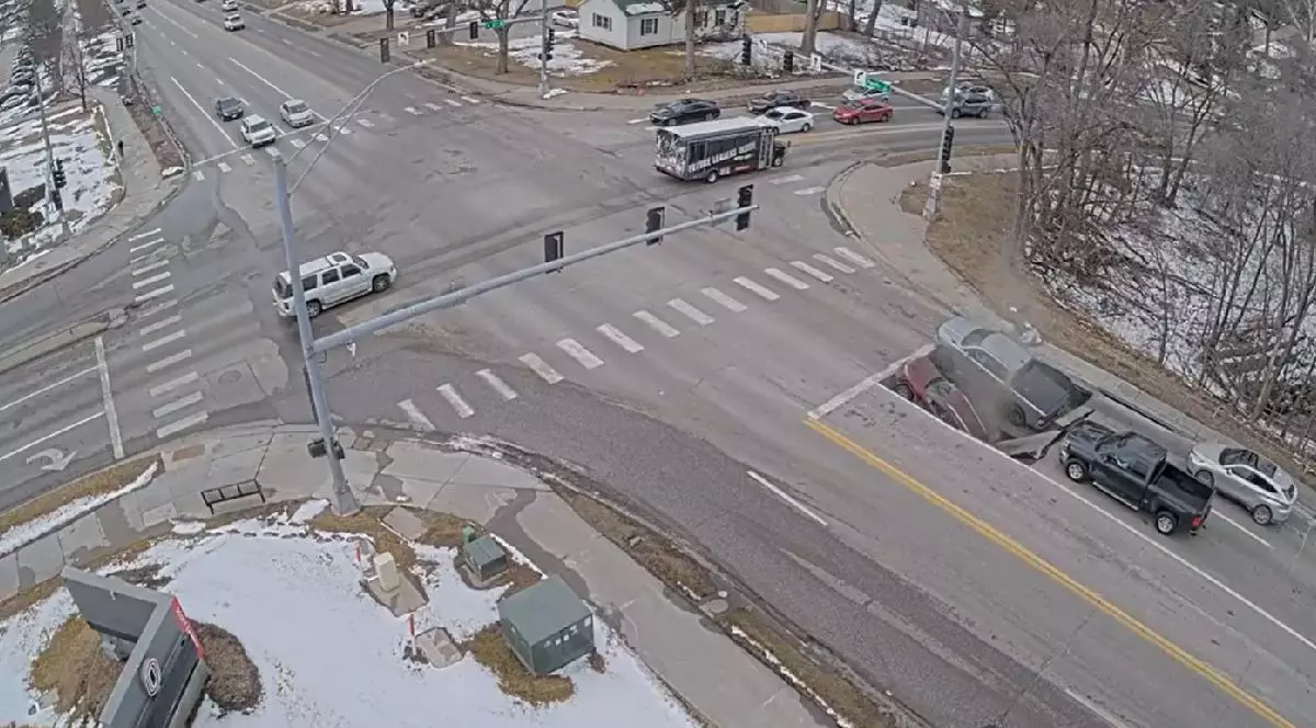 Imaginea arată o intersecție în timpul iernii. Drumul este în mare parte curat, cu petice de zăpadă și gheață. Un autobuz alb și mai multe mașini sunt oprite la trecerea de pietoni, așteptând la semafor.