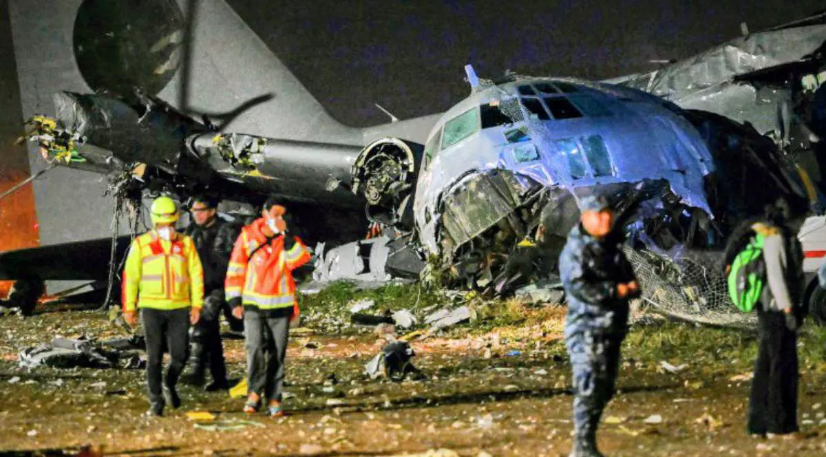 Un avion militar grav avariat, rupt în bucăți, stă pe un teren lângă pistă, noaptea. În jur sunt împrăștiate resturi de metal. Salvatori și polițiști, unii cu veste reflectorizante, merg printre epavă sub lumina reflectoarelor.