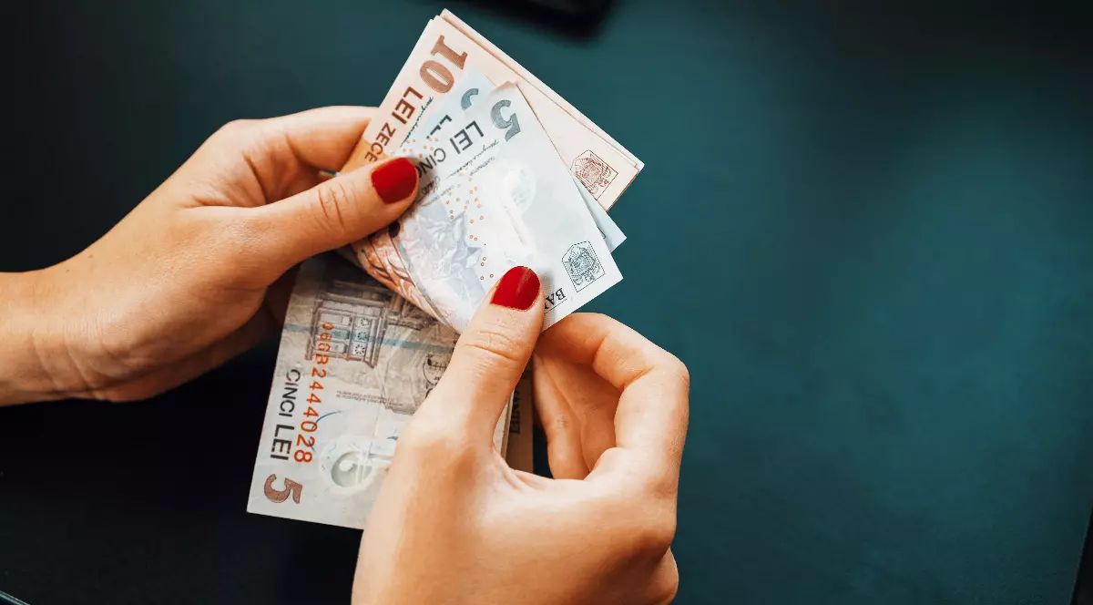 Imaginea surprinde o persoană care ține în mâini mai multe bancnote de lei românești, pregătindu-se să le numere. Se disting cupiuri de 5 și 10 lei, iar fundalul este unul simplu, de culoare închisă.