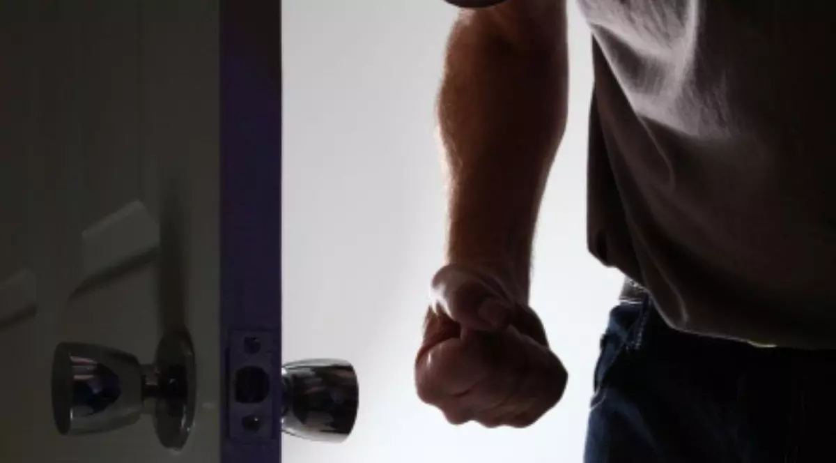 Bărbat agresiv intră într-o cameră întunecată, cu pumnul strâns. Fața nu este afișată.