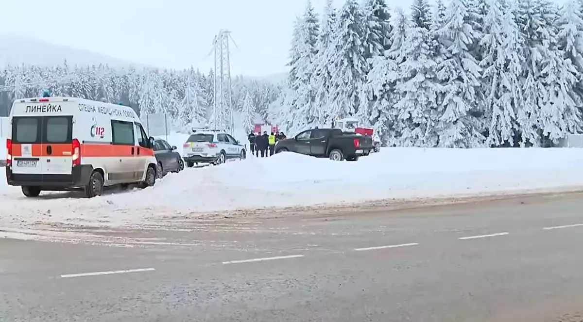 Fotografie cu un drum acoperit de zăpadă, pe care sunt parcate mai multe vehicule de urgență, inclusiv o ambulanță, lângă o zonă împădurită acoperită de zăpadă.