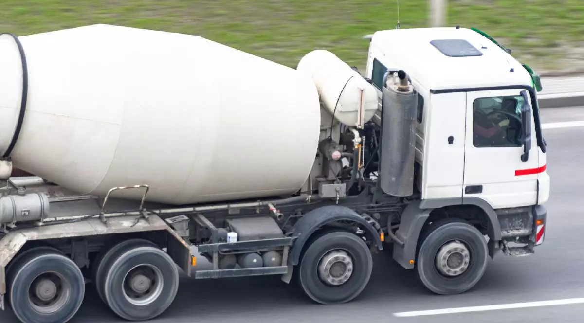 Un camion cu malaxor de ciment alb circulă pe o autostradă cu iarbă verde și mișcare neclară în fundal.