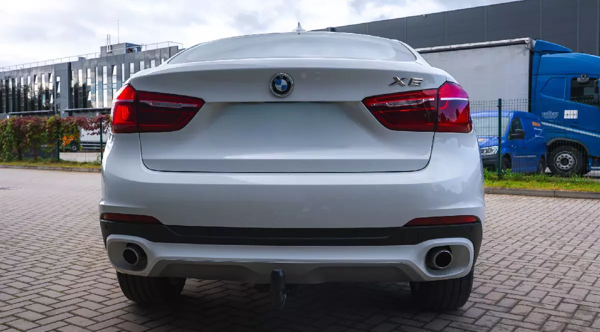 Fotografie a unui SUV BMW X5 alb, fotografiat din spate pe o parcare pavată. În fundal se văd un camion albastru și o clădire gri, alături de vegetație și un gard.