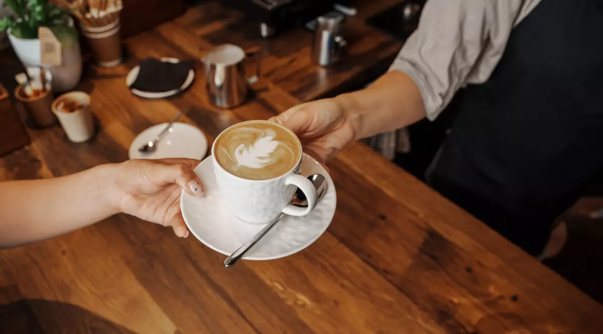 O ceașcă de latte cu un model în formă de frunză la suprafață. Cafeaua este servită într-o ceașcă albă, așezată pe o farfurioară asortată, alături de o linguriță metalică.