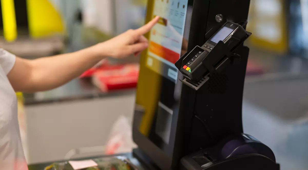 Imaginea surprinde o persoană care folosește o casă self-pay într-un supermarket, selectând opțiuni pe ecranul tactil în timp ce produsele sunt așezate pe bandă.