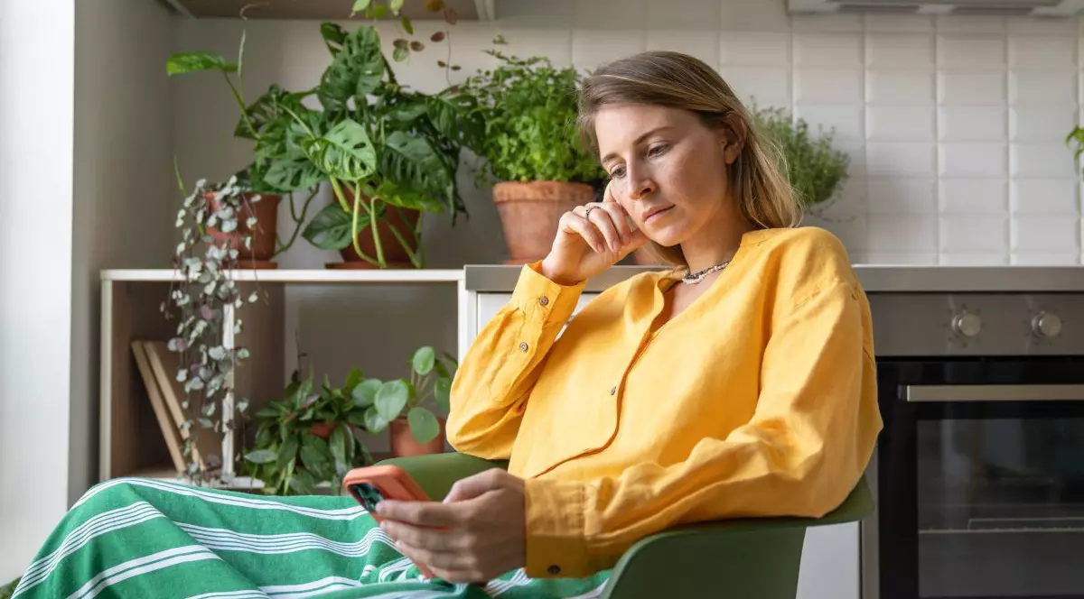 O femeie îmbrăcată într-o bluză galbenă se uită la telefon, stând jos.