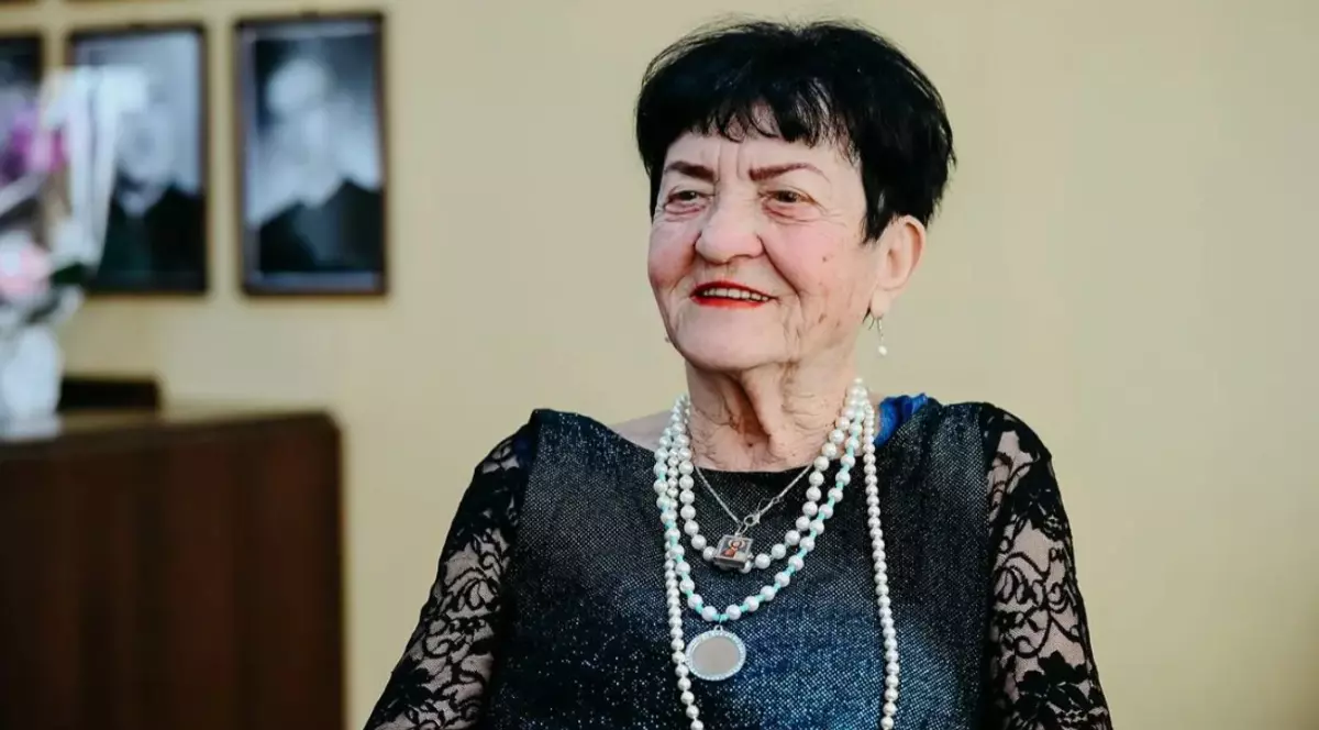 O femeie în vârstă de 90 de ani, brunetă cu părul tuns scurt și o rochie neagră din dantela, stă pe scaun și poartă la gât perle și o iconiță