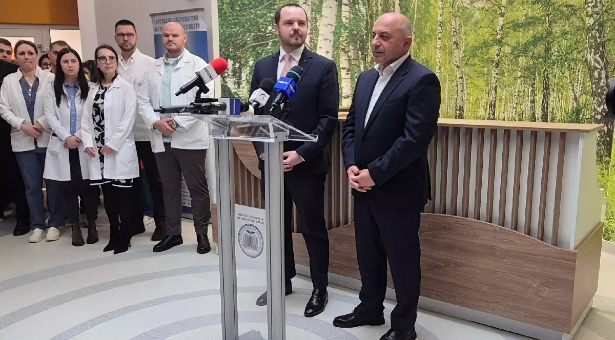 In noul Centru de Radioterapie Oncologica de la Spitalul Universitar de Urgenta Bucuresti sta la microfon in picioare un barbat impbracat in costum negru, este Catalin Cirstoiu, managerul spitalului. Langa el sta si il priveste un alt barbat, imbracat in costum negru, ministrul Sanatatii, Alexandru Rogobete. In fundal se vad oameni in halate albe.