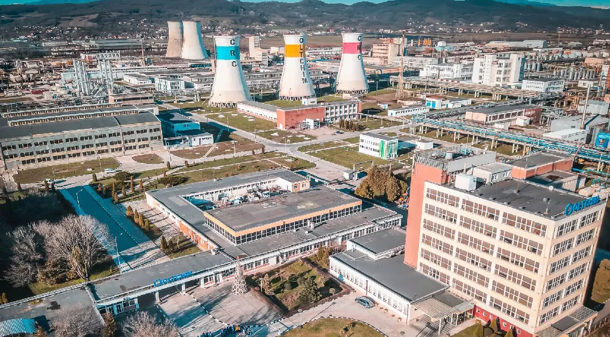 Imaginea prezintă o vedere aeriană amplă a unei platforme industriale. În plan îndepărtat, se remarcă mai multe turnuri de răcire masive. În prim-plan și în partea dreaptă se află clădiri de birouri.