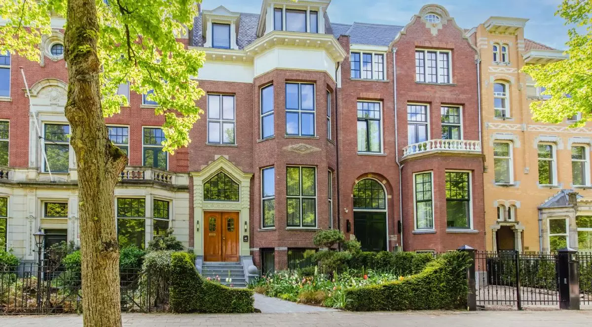 Fotografie cu o vilă istorică, din cărămidă, cu ferestre mari, fațade ornamentate și vegetație luxuriantă. Zi însorită, cu cer senin și copaci verzi, care încadrează scena.