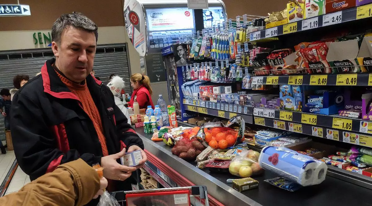 Fotografie cu un bărbat de vârstă mijlocie, cu părul scurt și cărunt, îmbrăcat într-o jachetă neagră cu roșu, care face cumpărături într-un magazin alimentar.