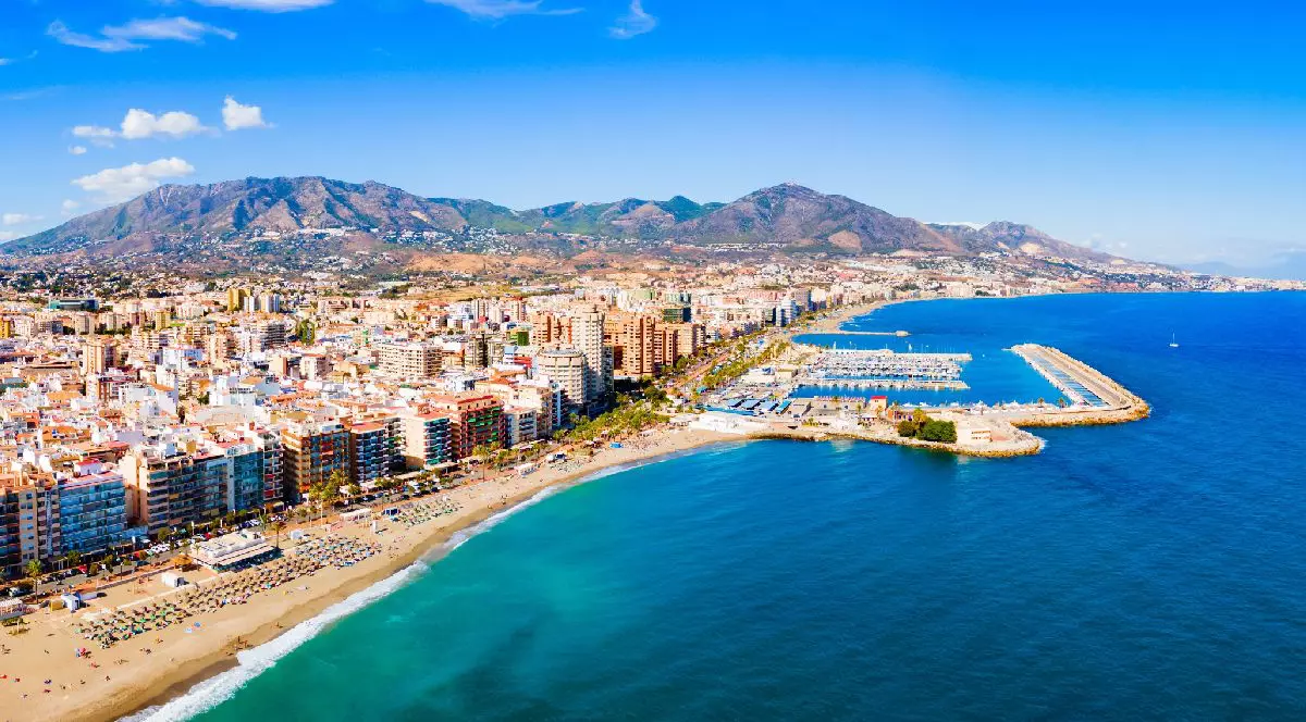 Fotografia surprinde o plajă largă, cu nisip auriu și apă turcoaz, pe care se află umbrele albe și șezlonguri. Clădiri înalte domină partea stângă, în timp ce un port cu numeroase iahturi ocupă partea dreaptă. Un dig se întinde în marea albastră, cu munți îndepărtați și accidentați sub un cer senin.