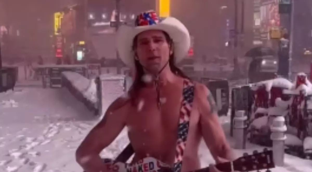 Un bărbat care este dezbrăcat și cântă la chitară pe o stradă unde ninge. Acesta poartă o pălărie pe cap.