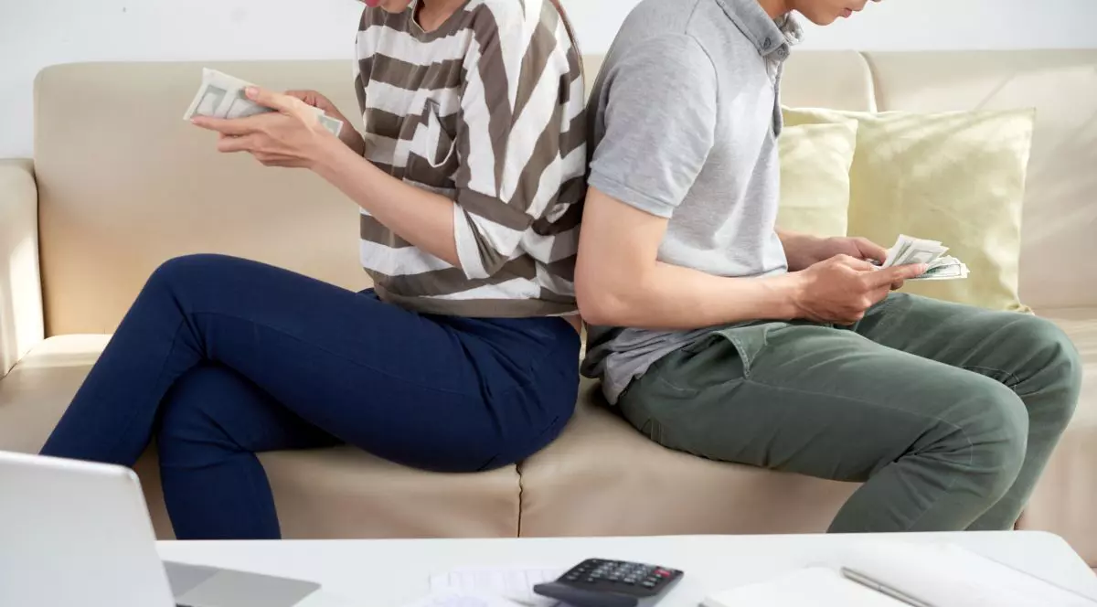 Unrecognizable young woman and man sitting back to back on cozy sofa and counting money, coffee table with bills on foreground