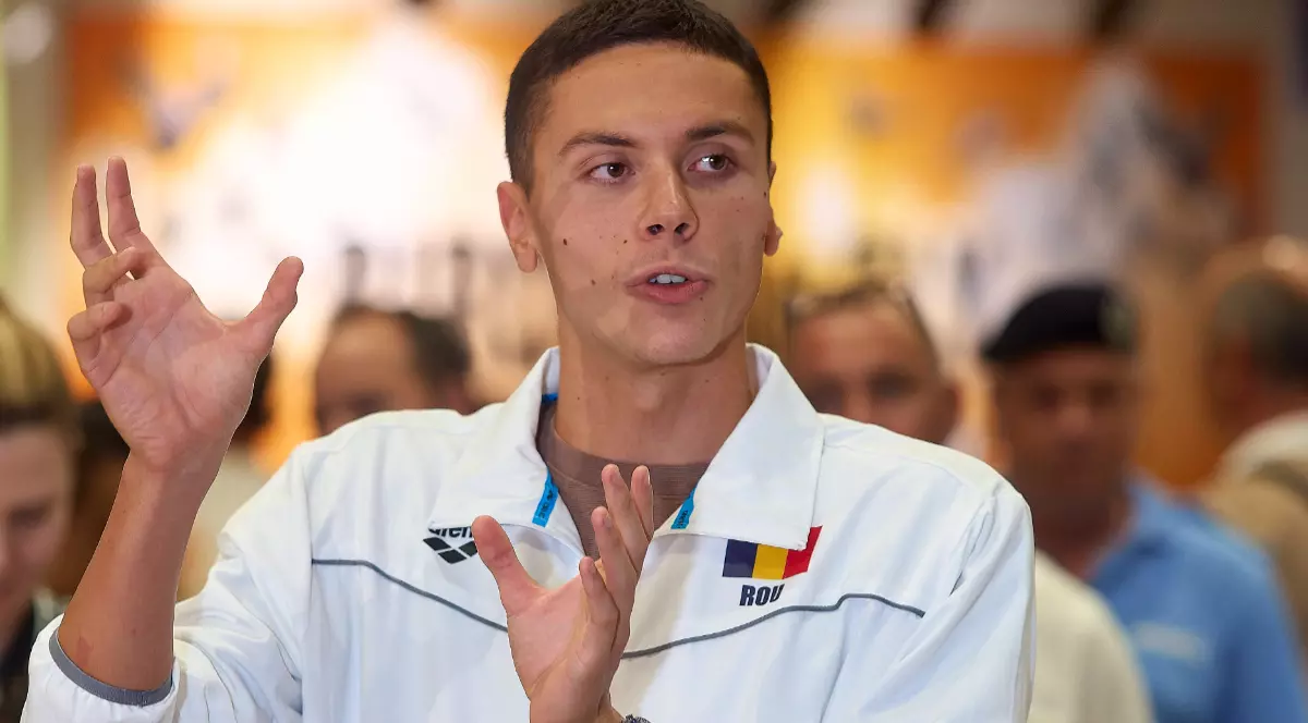 David Popovici, în timpul unei conferințe de presă la Aeroportul Otopeni, poartă un hanorac alb și gesticulează cu ambele mâini