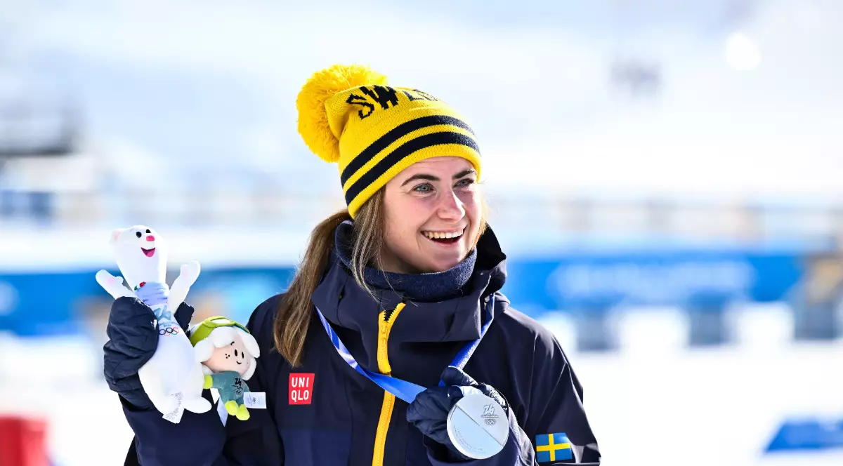 O sportivă zâmbește pe podium, purtând o medalie de argint la gât și o ținută de iarnă în culorile Suediei. În mâini ține mascotele competiției, iar atmosfera este una festivă, pe un fundal acoperit de zăpadă.