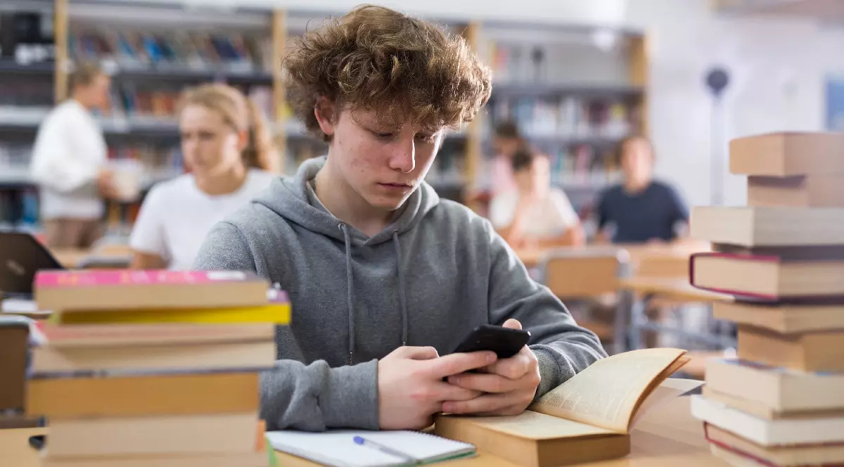 Portretul unui elev de cincisprezece ani stând la un birou din biblioteca școlii, ținând un telefon mobil în mâini