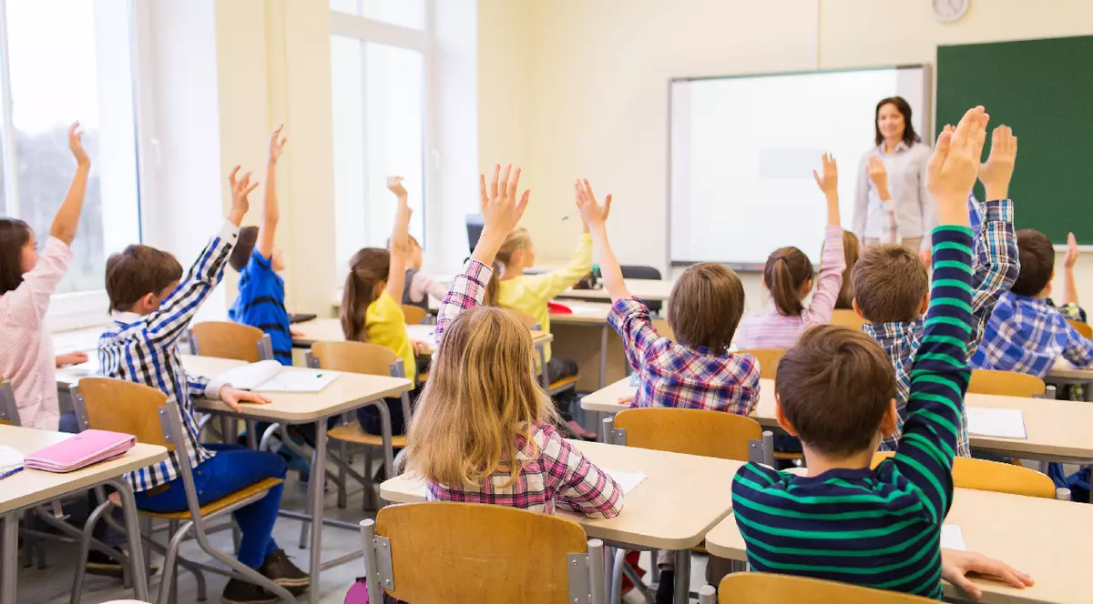 Mai mulți elevi, care stau în bănci, într-o sală de clasă, sunt cu mâinile ridicate