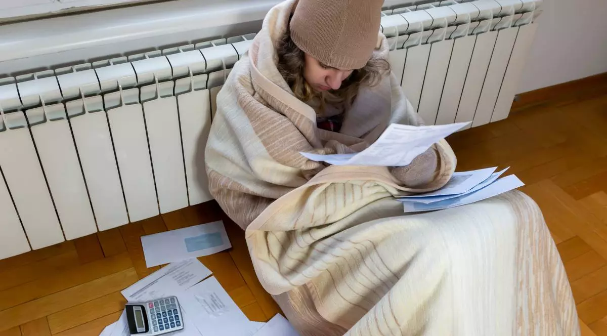 Tânără îngrijorată, invelita cu o patura crem, cu caciula pe cap, sta pe jos in casa. la geam, lipta de calorifer, citește factura de încălzire. Este confuză și uimită de vești incredibile: factură fiscală mare, notificare de datorie, raport financiar prost, probleme cu banii