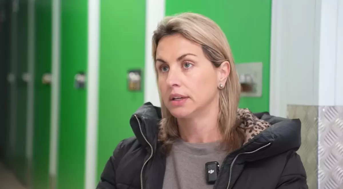 O femeie blondă cu haină neagră, în timp de dă un interviu, în spatele ei se văd dulapuri cu uși verzi