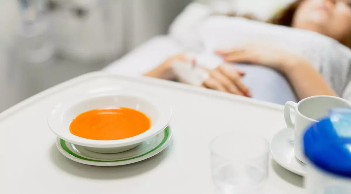O femeie care stă întinsă în pat, în spital, are branulă în mână și pe masuță are o farfurie de ciorbă