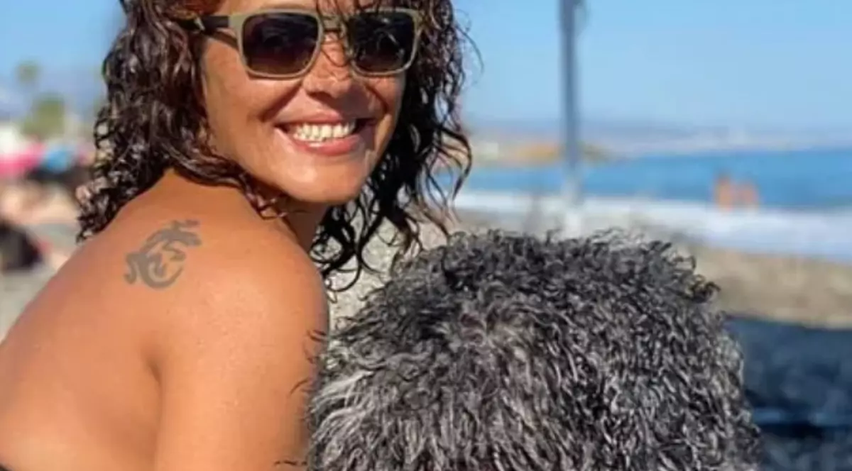 O femeie cu părul creț și ochelari de soare la ochi, pe o plajă împreună cu cățelușa ei