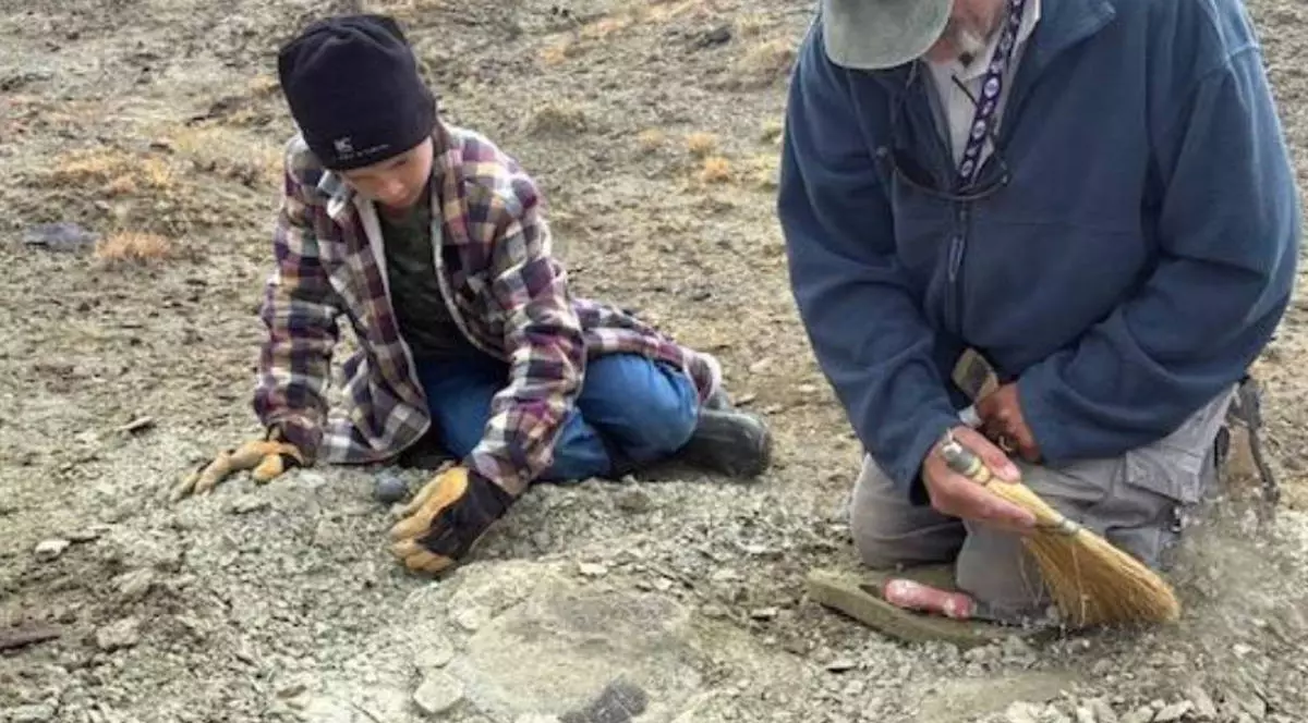 Touren Pope, în vârstă de unsprezece ani, in camasa in carouri, caciula neagra, asezat pe jos, ajuta la excavare alături de JP Cavigelli, specialist în colecții muzeale la Muzeul Geologic Tate de la Colegiul Casper din Wyoming, asezat in genunchi, cu un hanorac albastru inchis si o sapca pe cap, cu o maturica in mana, Fotografie de Craig Thomas, Biroul Local BLM Rock Springs