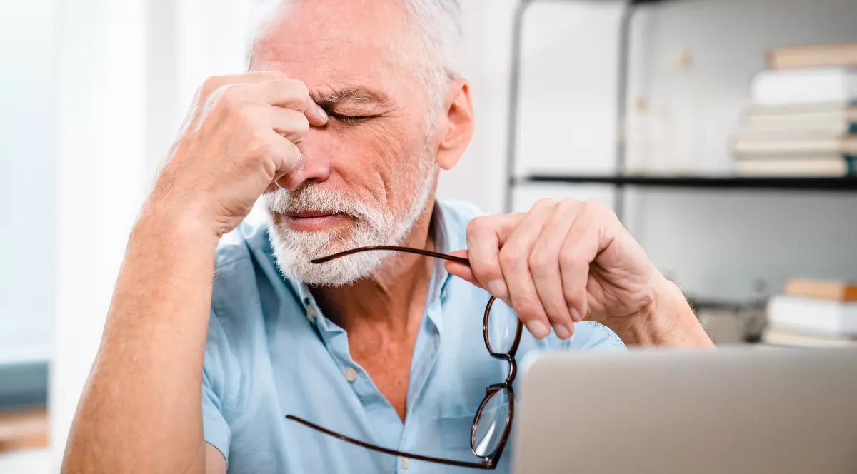 Un bărbat în vârstă stă la birou, ținând ochelarii în mână și apăsându-și fruntea cu degetele, ca și cum ar fi obosit sau îngrijorat. În fața lui se află un laptop, iar expresia feței sugerează stres sau concentrare intensă.