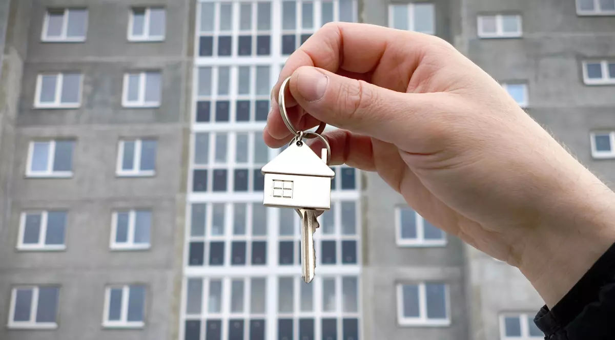 O mână care ține o cheie cu un breloc cu o casă. Pe fundal se vede un bloc vechi.