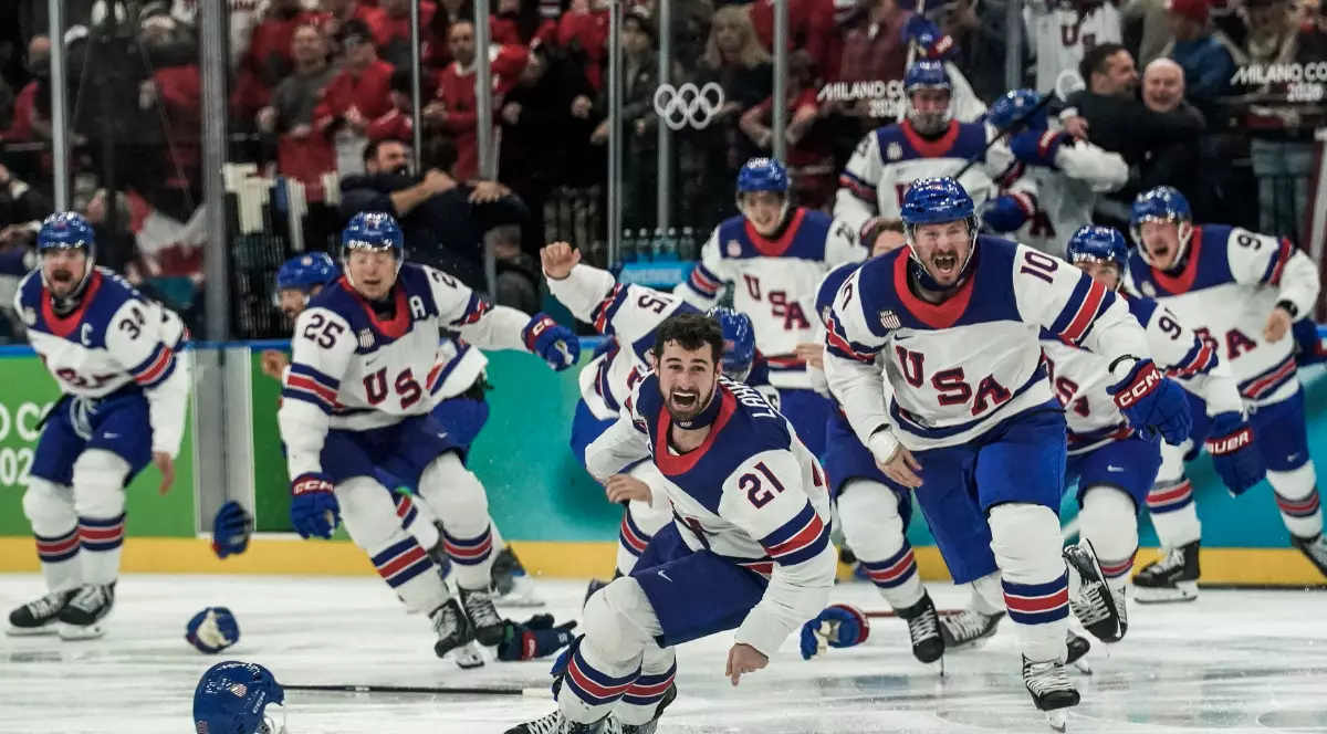 Jucătorii echipei SUA sărbătoresc exploziv pe gheață după golul decisiv, aruncându-și echipamentul în aer și alergând unii spre alții într-un moment de euforie totală. În fundal, tribunele sunt în delir, iar atmosfera reflectă un triumf istoric obținut într-o finală dramatică.