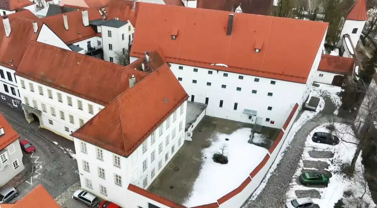 Fotografia surprinde o vedere aeriană a unui castel istoric, cu ziduri albe și acoperiș din țiglă roșie. Arhitectura se caracterizează prin liniile sale dreptunghiulare și ferestrele mici, dispuse uniform. Adiacent se află o curte acoperită de zăpadă și câteva vehicule parcate în partea dreaptă.