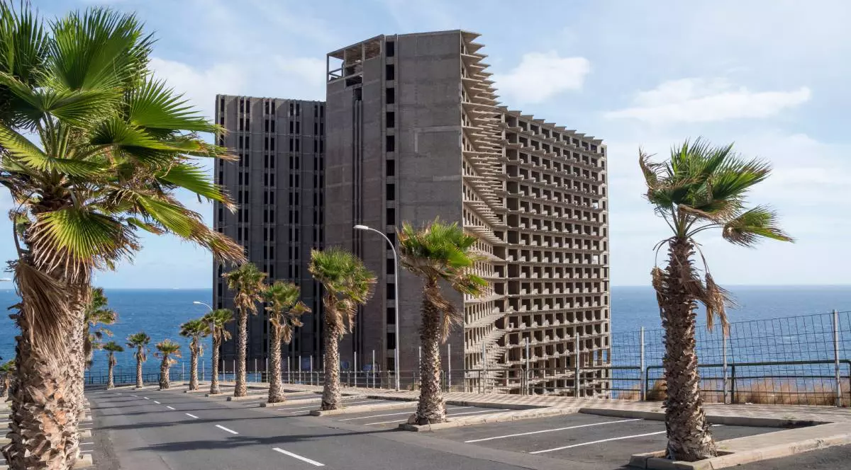 Hotelul Añaza, abandonat și neterminat - Acoran, Tenerife, Spania, se vede constructia, in departare marea, palmieri in prim plan pe marginea unui drum, cerul este senin si este o zi insorita