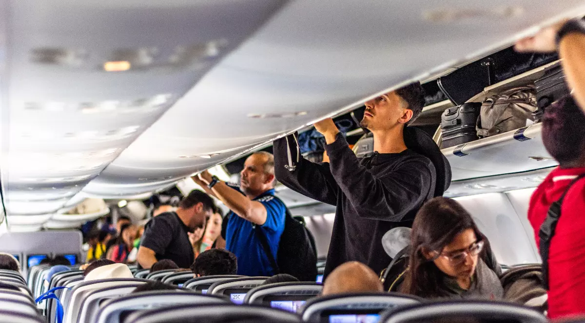 Pasagerii se află în avion în timpul îmbarcării, încercând să își așeze bagajele în compartimentele de deasupra scaunelor. Cabina este aglomerată, iar oamenii stau în picioare pe culoar, în timp ce alții sunt deja așezați pe locurile lor.