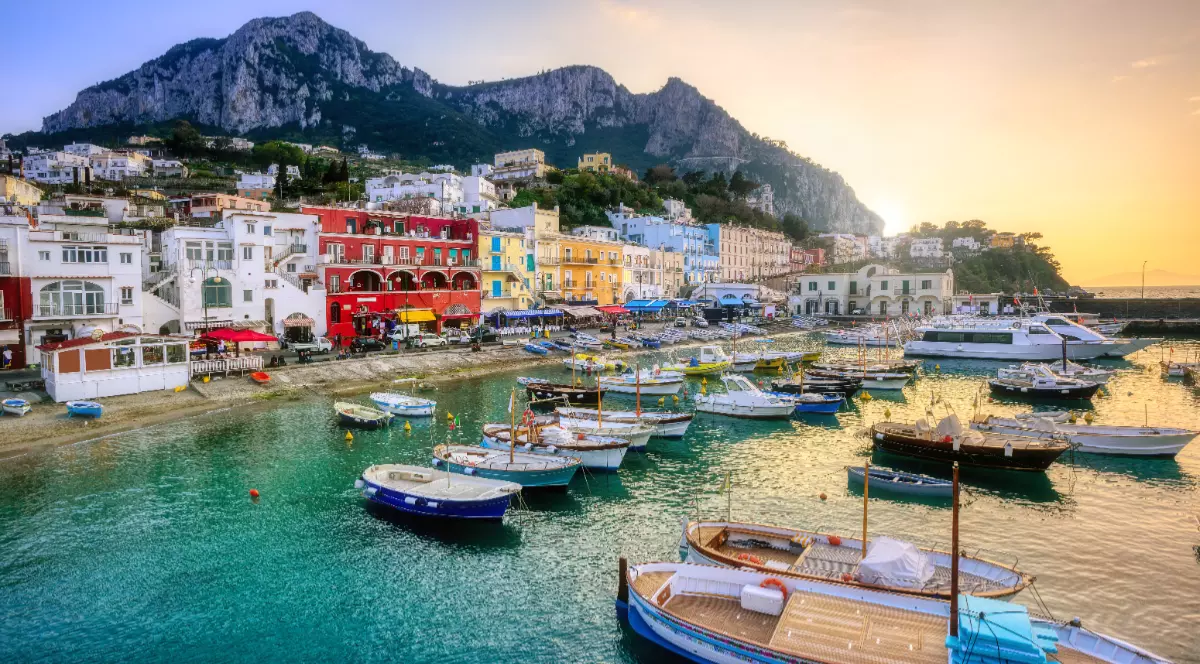Insula Capri din Italia, bărcile sunt în port, soarele apune în spatele muntelui stâncos, iar casele colorate de la malul mării sunt admirate de turiști