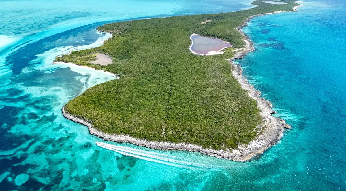 Fotografie aeriană a unei insule luxuriante și verzi, cu o lagună centrală, înconjurată de ape turcoaz și recife de corali, cu o barcă albă navigând în apropiere.