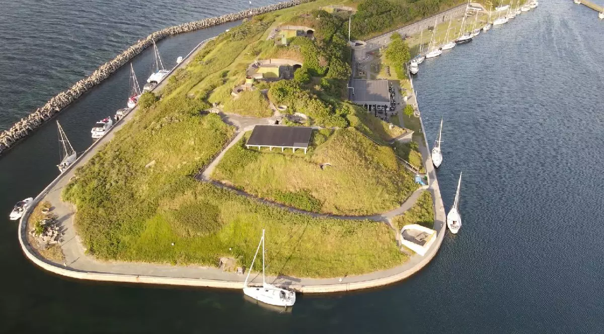 Fotografie aeriană a unei mici insule acoperite cu iarbă, cu câteva clădiri, înconjurată de un port cu bărci cu pânze acostate și un dig stâncos în partea stângă.