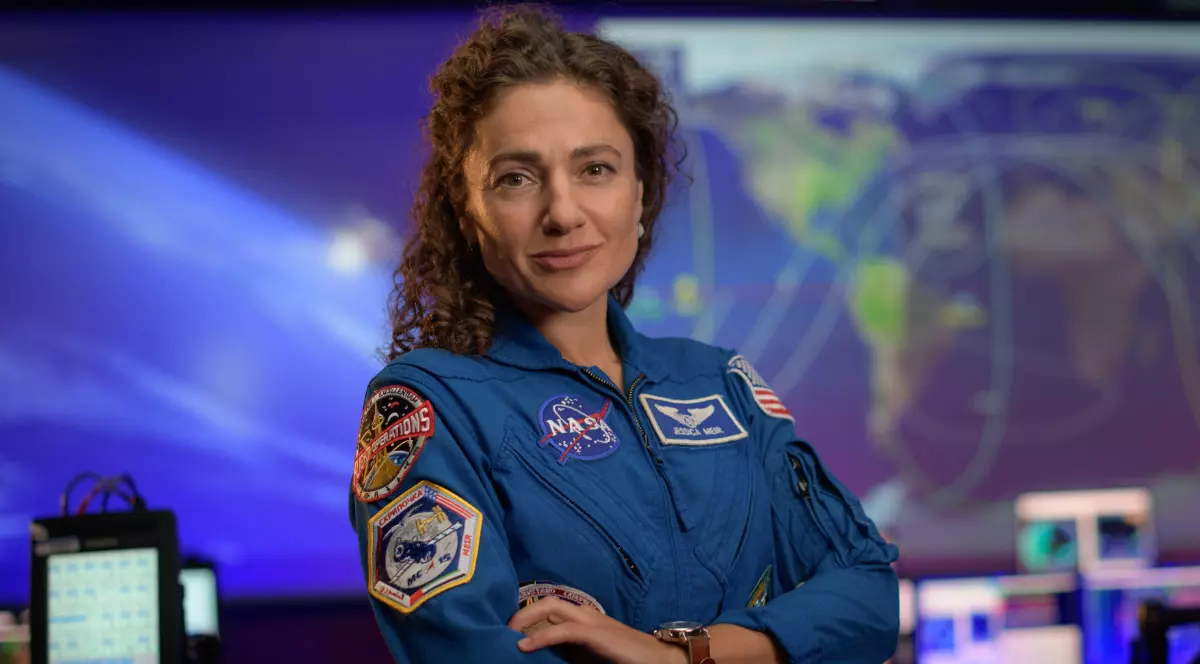 O astronaută poartă un costum NASA și stă cu brațele încrucișate, într-un centru de control cu ecrane și hărți în fundal.