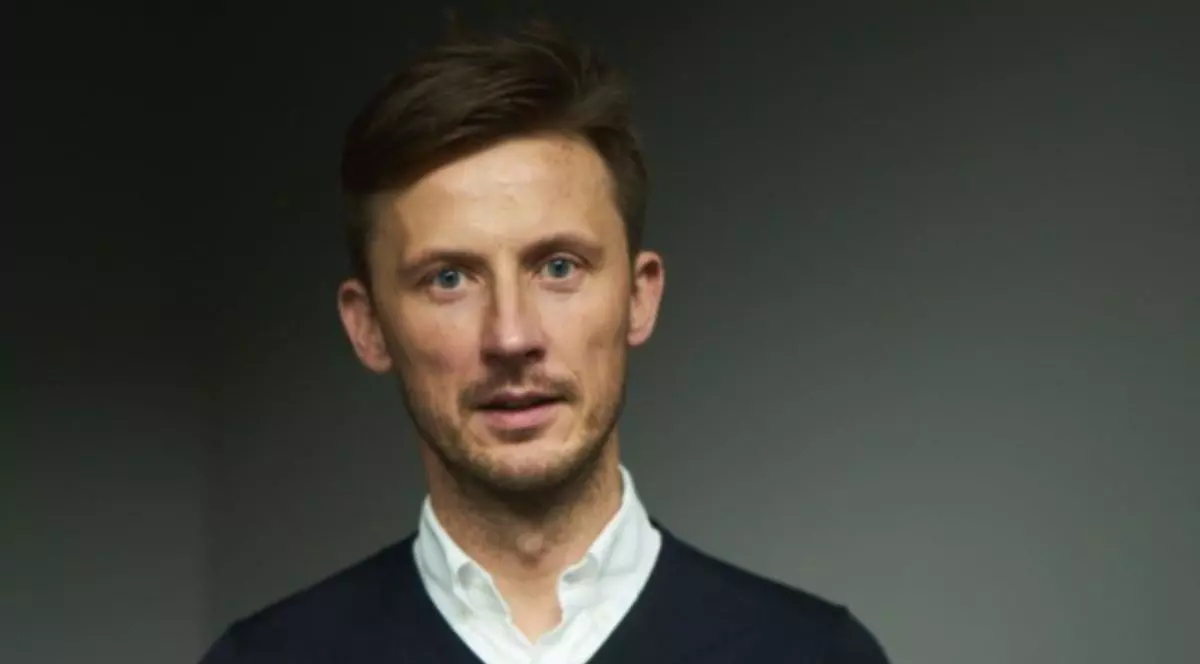 Krystian Wolak, pe fundal neutru, îmbrăcat cu o cămașă albă și un pulover negru pe deasupra