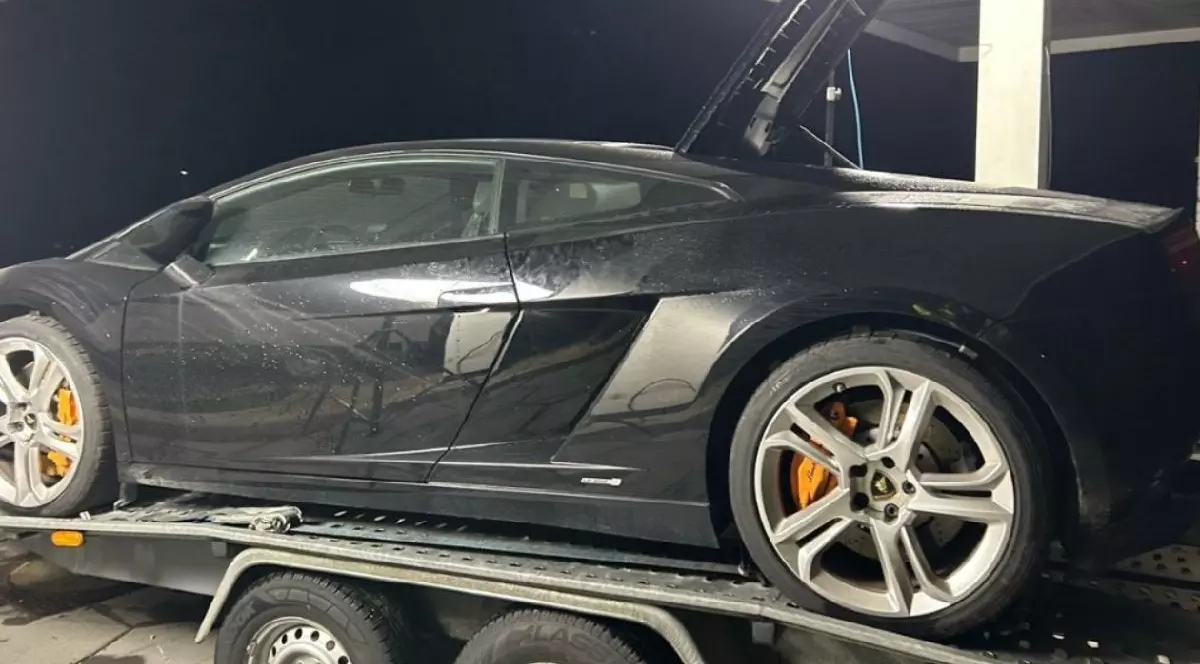 Imaginea prezintă un Lamborghini negru așezat pe o platformă. Mașina are jante mari din aliaj argintiu. Fundalul este întunecat.