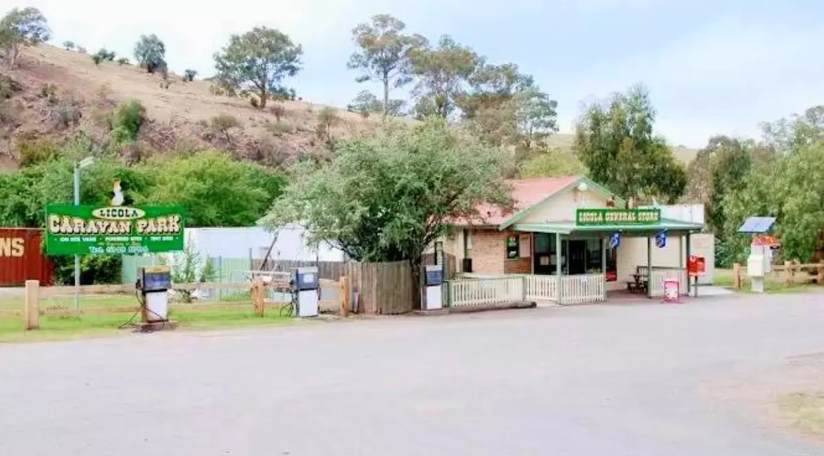 Strada din ora;ul australian Licola, se vede o strada larga, in spate un magazin cu un banner verde la intrare si acoperis maro, copaci in jur și in depărtare, nu este nimeni pe stradă