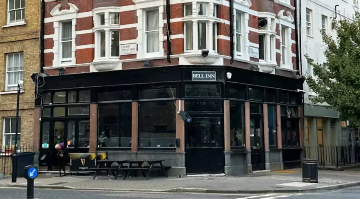 Imaginea surprinde o clădire de colț din Londra, cu fațadă din cărămidă roșie și elemente decorative albe, la parterul căreia se află un pub numit „Bell Inn”. În față se văd mese de exterior și trotuarul unei intersecții tipice din centrul orașului.