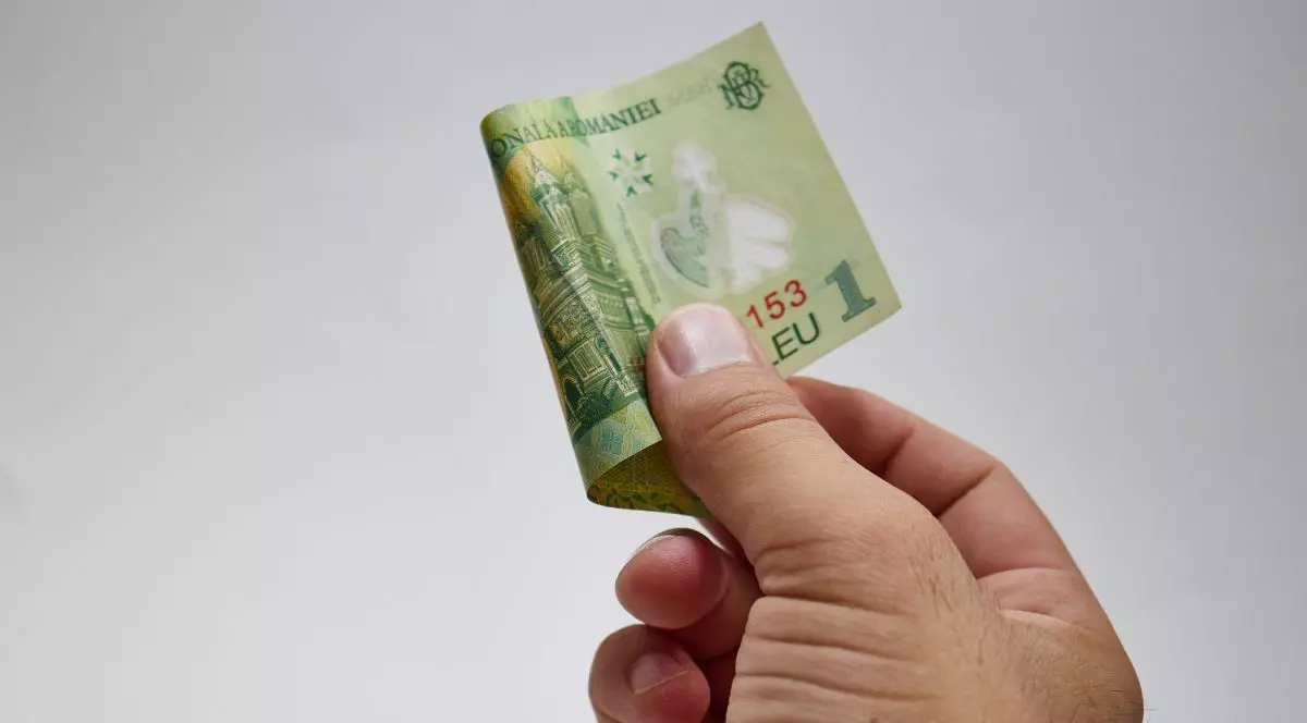 Mâna unui bărbat ține cu două degete o bancnotă de un leu, pe fundal gri neutru